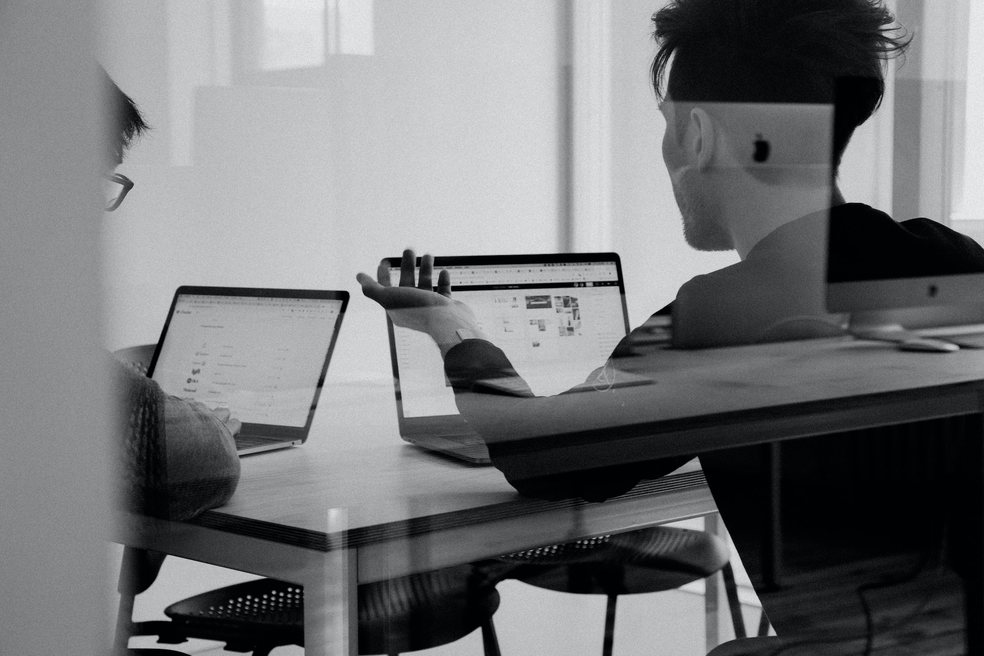 Black and white photo looking through a conference room glass wall at two individuals having a discussion and working on their laptops, representing business ownership disputes